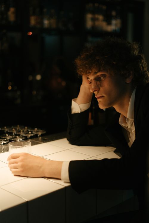 Free A young man in a black suit looking pensive while seated at a bar counter with a drink. Stock Photo