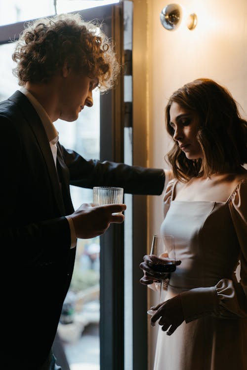 Man in Black Suit Jacket Holding Drinking Glass Beside Woman in White Dress