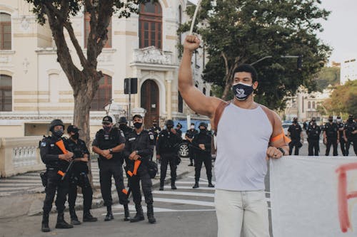 Protesting Man in White Tank Top and White Pants Standing on the Street Near a Group pf Policemen