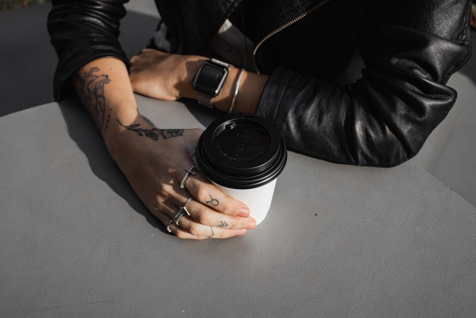 A tattooed person in black leather jacket holds a coffee cup indoors, showing rings and smartwatch.