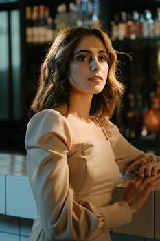 A stylish woman in a beige dress sits elegantly at a bar counter in a dimly lit restaurant, gazing thoughtfully.