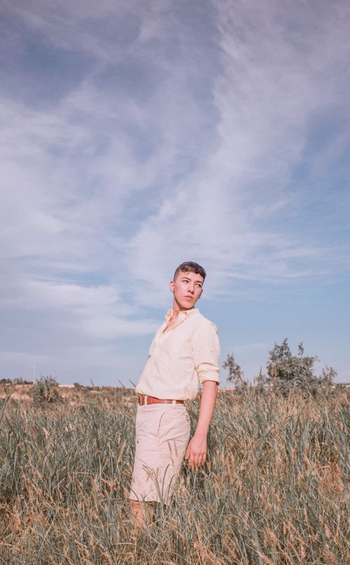 Free A young man posing confidently in a sunlit grassy field with a clear blue sky. Stock Photo