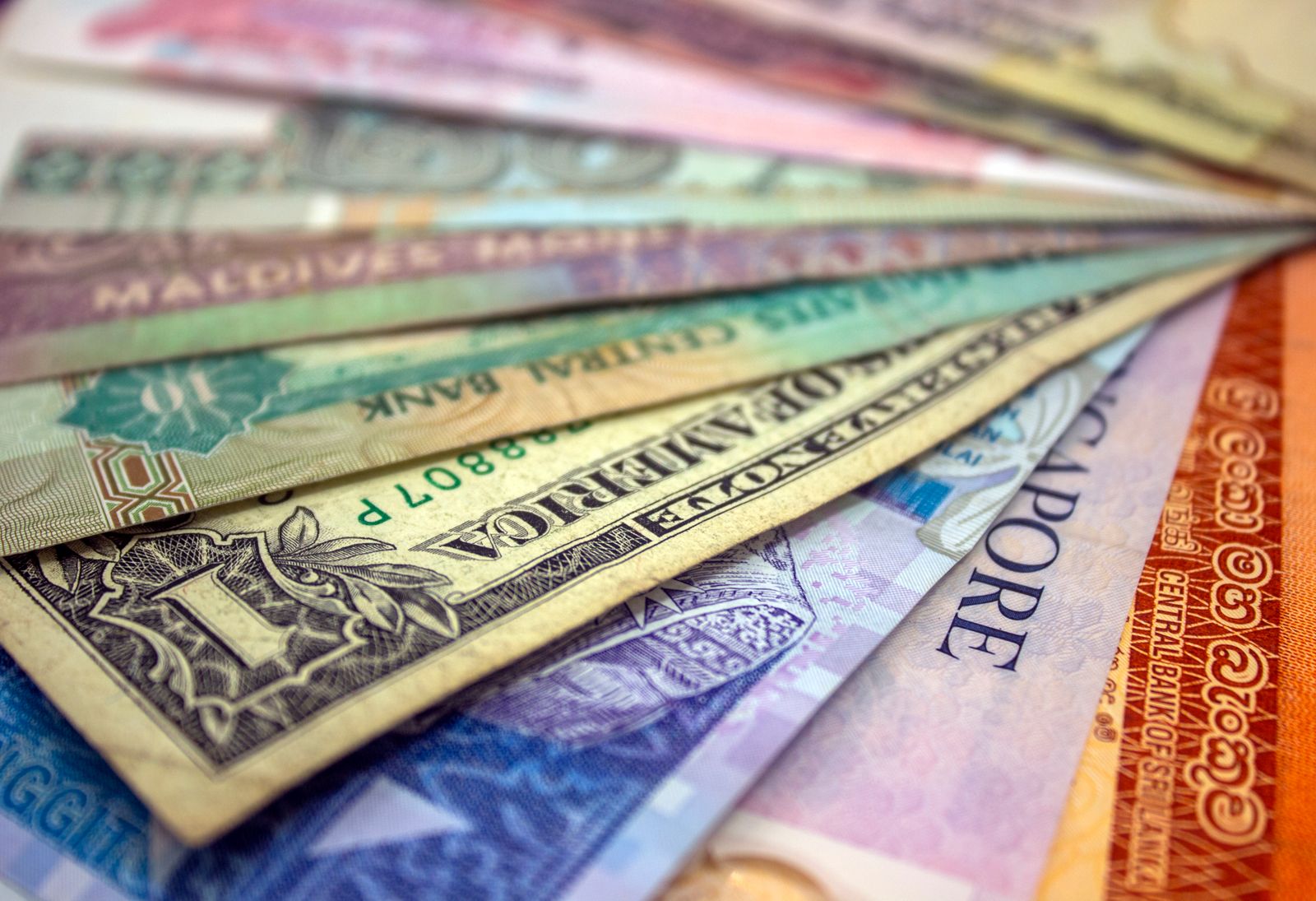A fanned stack of international banknotes from different countries, including Maldives, Singapore and Sri Lanka currencies, alongside a U.S. one-dollar bill