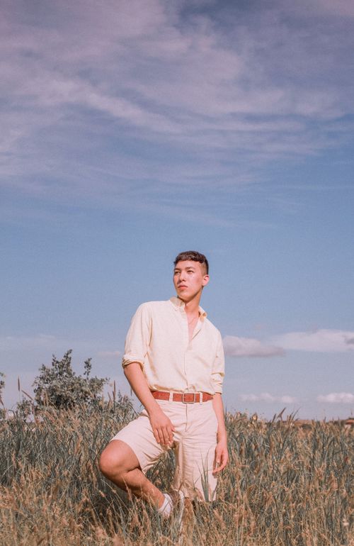 Free Young man in stylish attire poses thoughtfully in a grassy field under a blue sky. Stock Photo