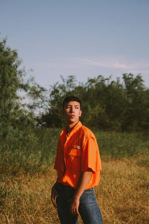 Free Pensive young man in an orange shirt posing outdoors in a sunlit field. Stock Photo
