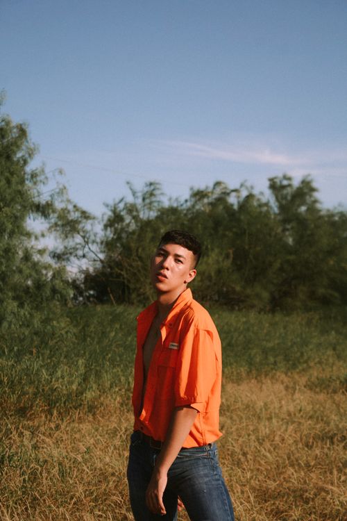 Free A young man poses confidently in a grassy field with an orange shirt under a clear blue sky. Stock Photo