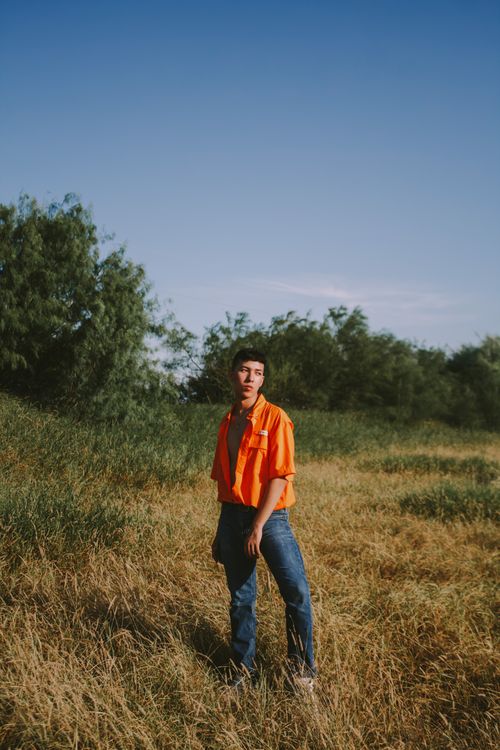 Free A young man in an orange shirt stands in a grassy field under a blue sky, exuding a contemplative mood. Stock Photo