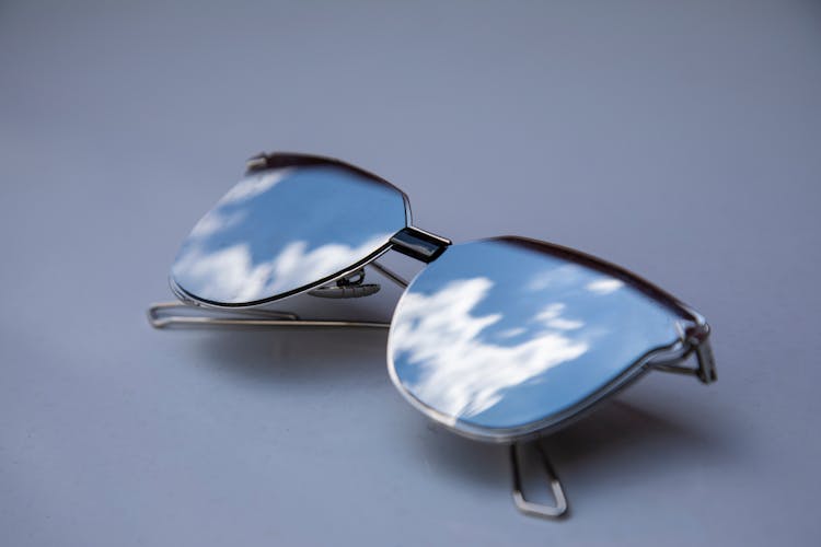 Silver Framed Sunglasses With Reflection Of Blue Sky And White Clouds