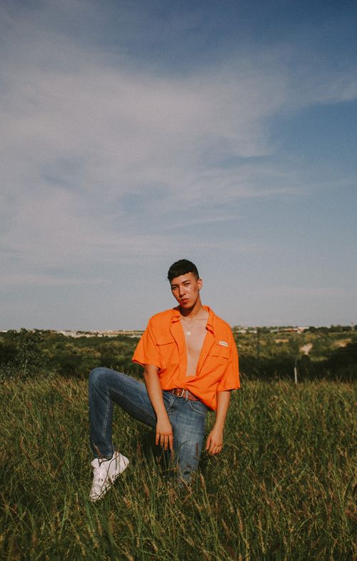 Free Stylish young man in vivid orange shirt kneeling in a green field under a clear blue sky, showcasing modern fashion. Stock Photo