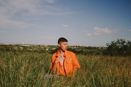 Free Man wearing orange posing thoughtfully in a lush grass field under blue sky. Stock Photo