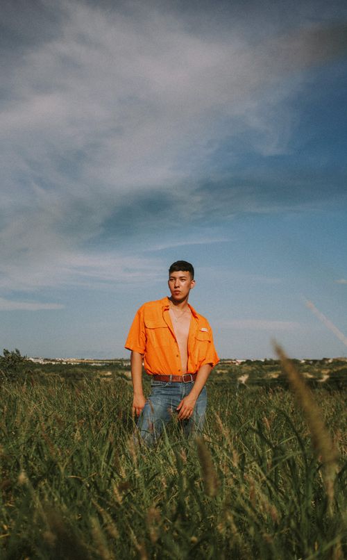 Free Adult male posing in a grassy field wearing an orange shirt under a blue sky. Stock Photo