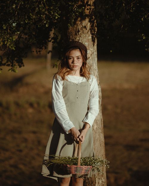 Free Pretty female in vintage outfit standing in park near tree holding basket full of flowers and looking at camera Stock Photo