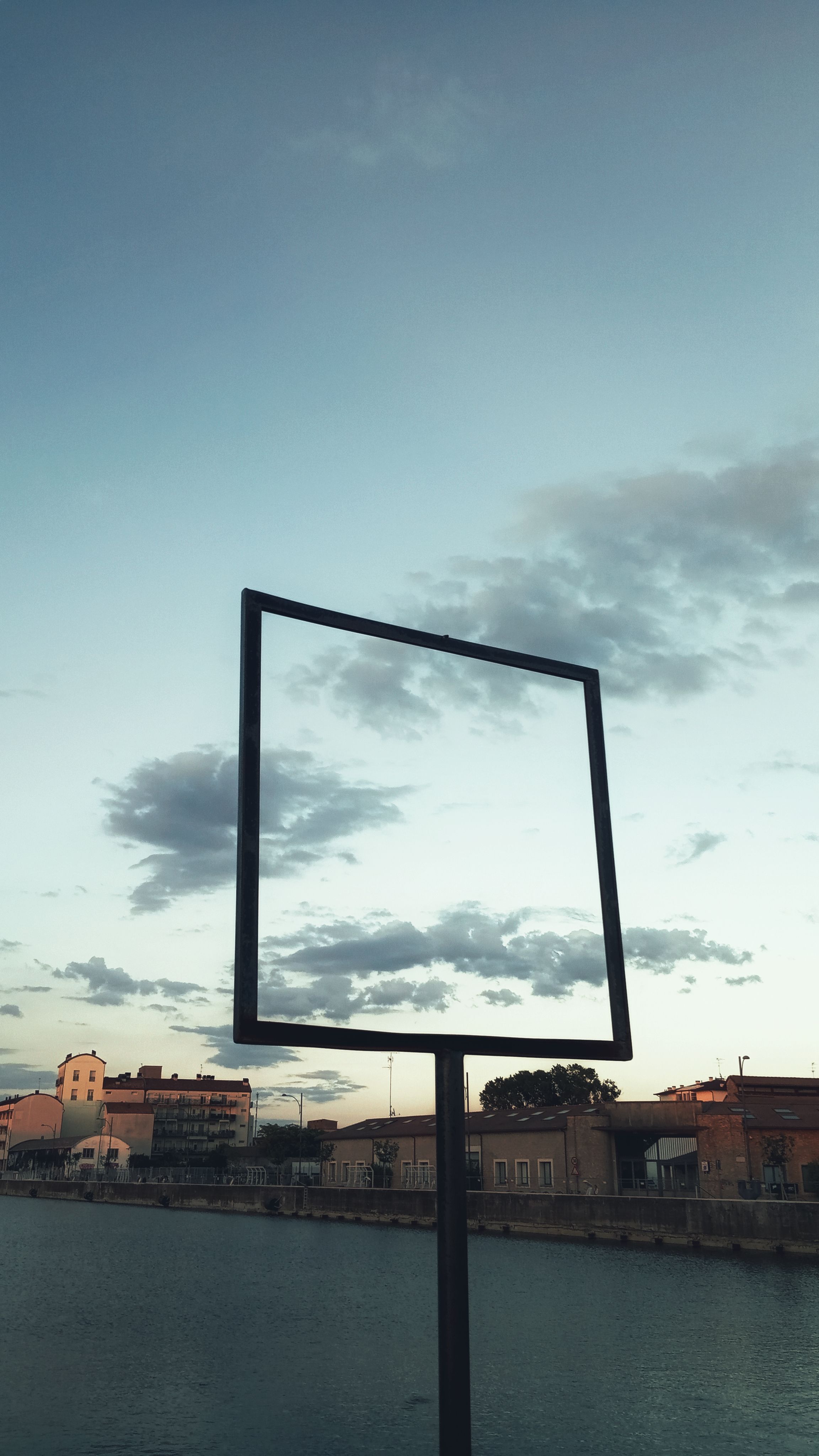 Empty billboard standing on pillar near river and industrial buildings under blue cloudy sky