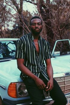 Confident African American man in glasses and striped shirt sitting on vintage car at daytime