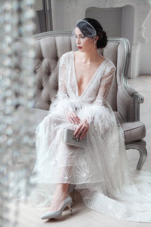 Full length calm graceful bride wearing luxurious white gown sitting on comfy armchair in studio