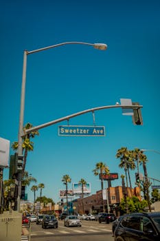 Busy city intersection on Sweetzer Ave in Los Angeles with palm trees and traffic under a clear blue sky.