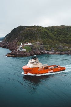 An orange cargo vessel navigates through calm sea waters near a rocky coastline with a lighthouse in view.