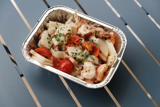 From above of tasty squids with tomatoes and herbs under sauce in foil container on blue metal table