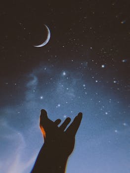 Silhouetted hand reaching out to a crescent moon amid a starry sky, creating a dreamy and celestial scene.