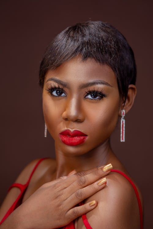 Young trendy African American female with accurate manicure in red dress looking at camera on brown background