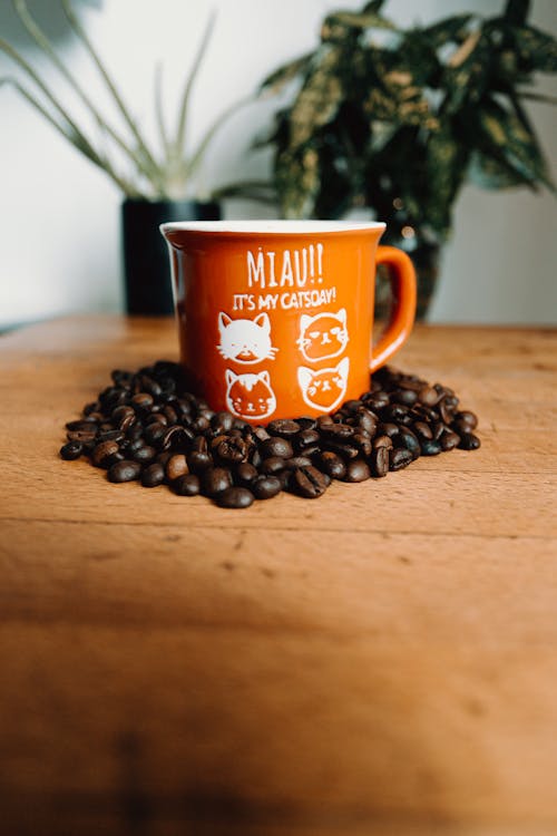 Ceramic orange mug with hot coffee placing on wooden table with roasted coffee beans around beverage