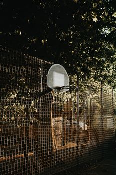 Old empty athletic field with basketball hoop on metal fence under tree in sun lights