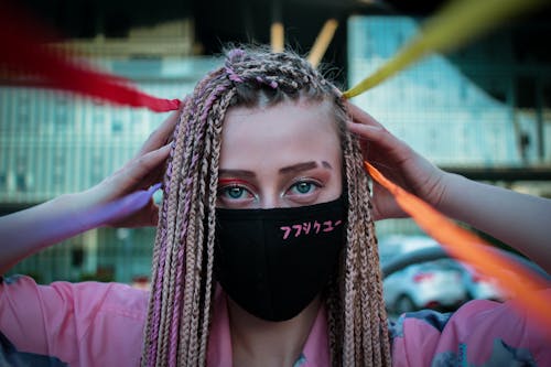 Free Cool young informal woman with long colorful braids wearing black safety mask looking at camera in city Stock Photo