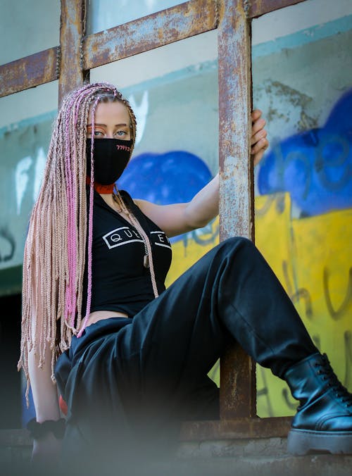 Free Full length of young female with long colorful braids wearing black outfit with medical mask sitting on grunge fence Stock Photo