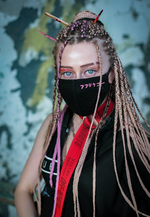 Free Cool modern woman with long braided hair wearing black mask and looking at camera against shabby wall Stock Photo