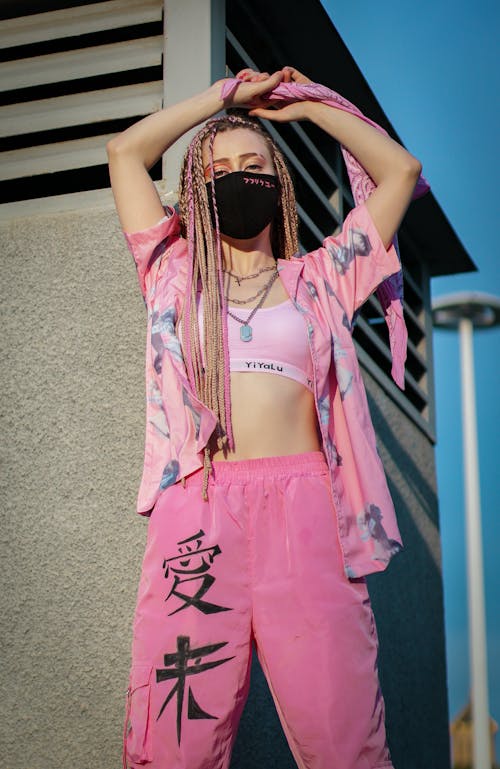 Low angle of cool woman in pink street style outfit and black medical mask raising arms and looking at camera