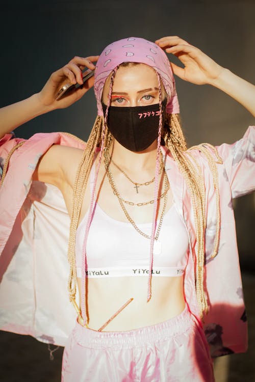 Cool contemporary female wearing pink top with shirt and headscarf with black safety mask looking at camera in sunlight