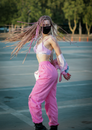一位穿着粉红色衣服、戴着面具的现代年轻女性，在色彩鲜艳的长辫飞舞中，于城市停车场上跳舞