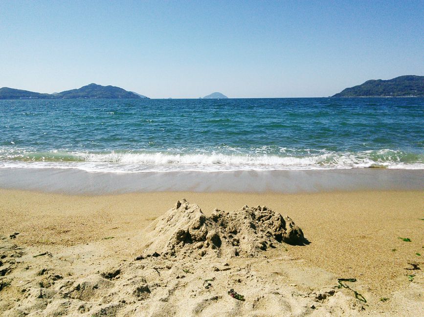 Tranquil beach scene with sand, ocean waves, and distant islands under a clear blue sky.