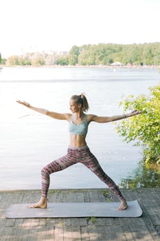 A woman in activewear performing yoga on a mat by a scenic lake in a lush, sunny setting.