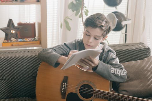 A young boy sits on a couch writing music in a notebook while holding an acoustic guitar.