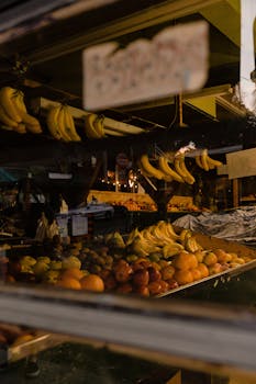 Colorful fruits on display at an evening farmers market stall with bananas, apples, and citrus fruits.