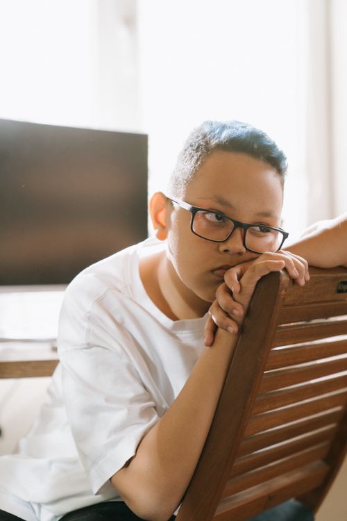 Free Young boy with glasses sitting indoors, leaning on chair, appearing thoughtful. Bright light fills the room. Stock Photo