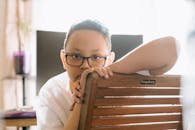 一个戴着眼镜的男孩正坐在室内的椅子上，若有所思