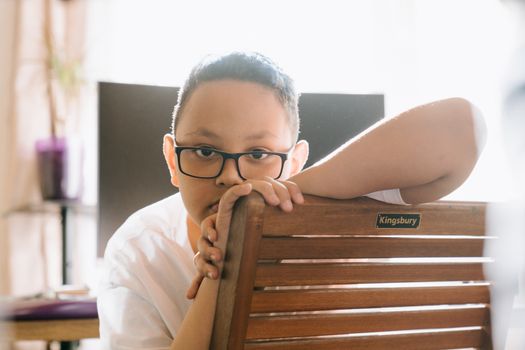 一个戴着眼镜的男孩正坐在室内的椅子上，若有所思