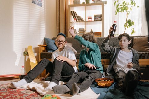 Free Three boys having fun with snacks and drinks, playing indoors with a skateboard and gadgets. Stock Photo