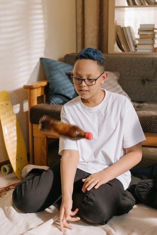 Free Young person with blue hair and glasses flips bottle indoors, having fun. Stock Photo