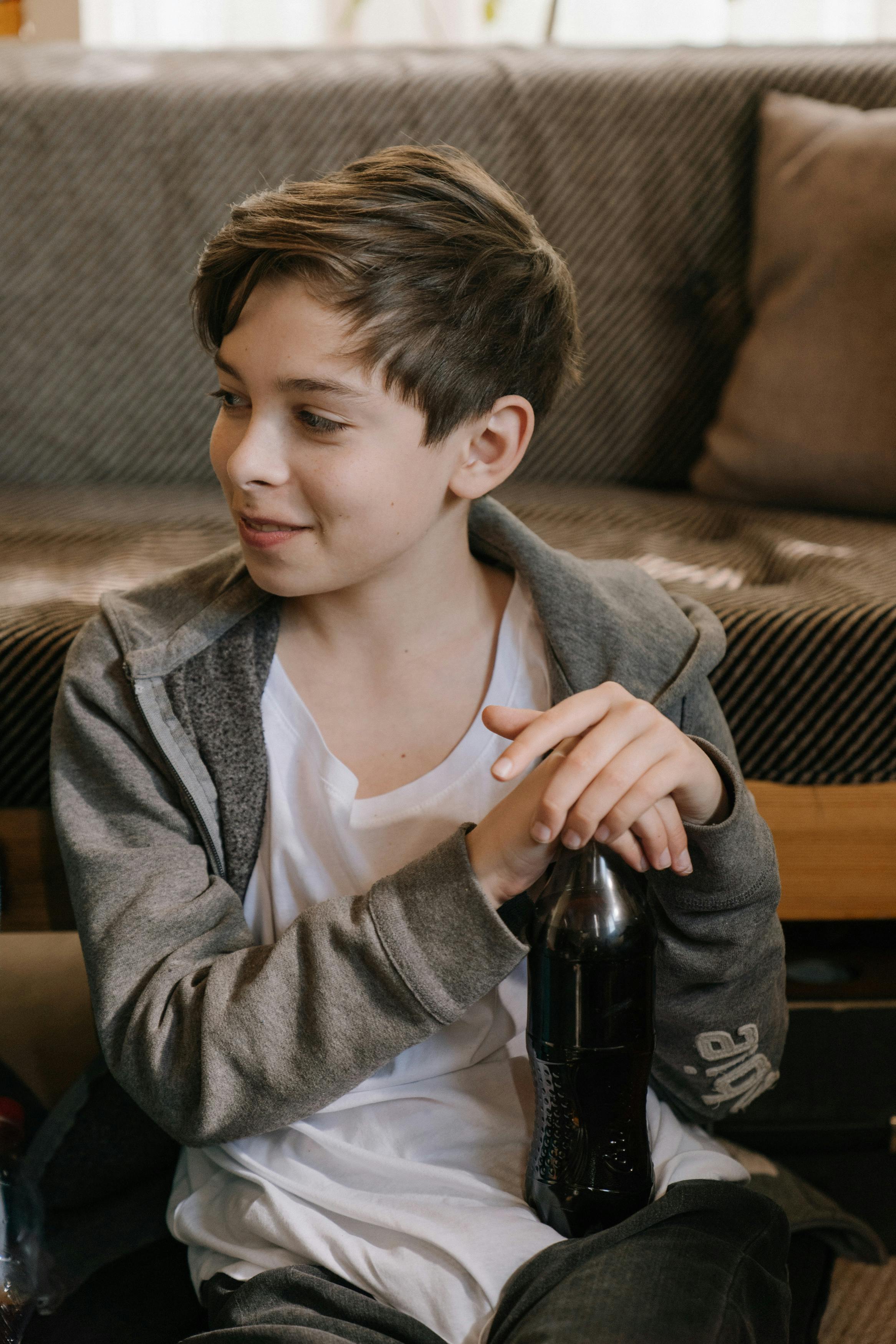 A young boy sits indoors, smiling while holding a soda bottle, wearing casual clothing.