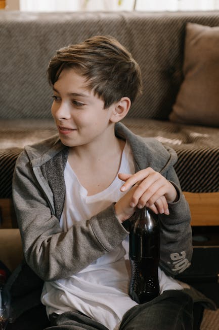 A young boy sits indoors, smiling while holding a soda bottle, wearing casual clothing.