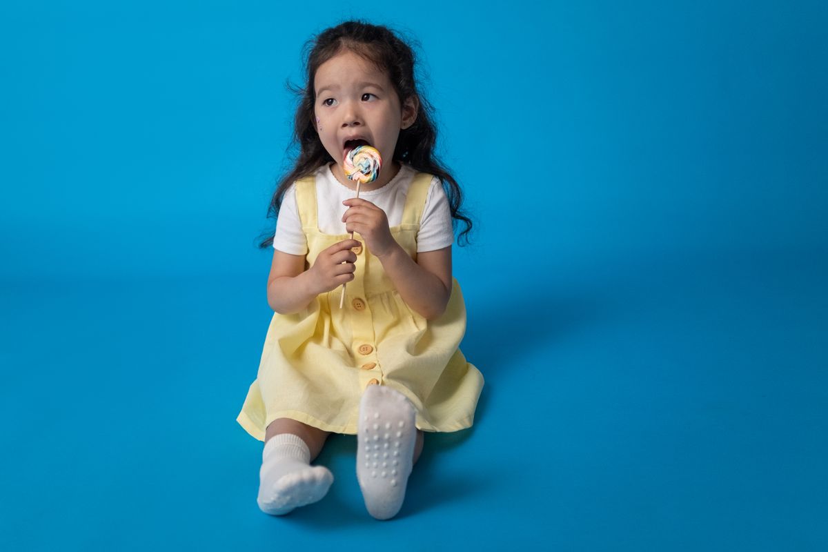 Adorable young girl in yellow dress enjoying a lollipop against a vibrant blue background.