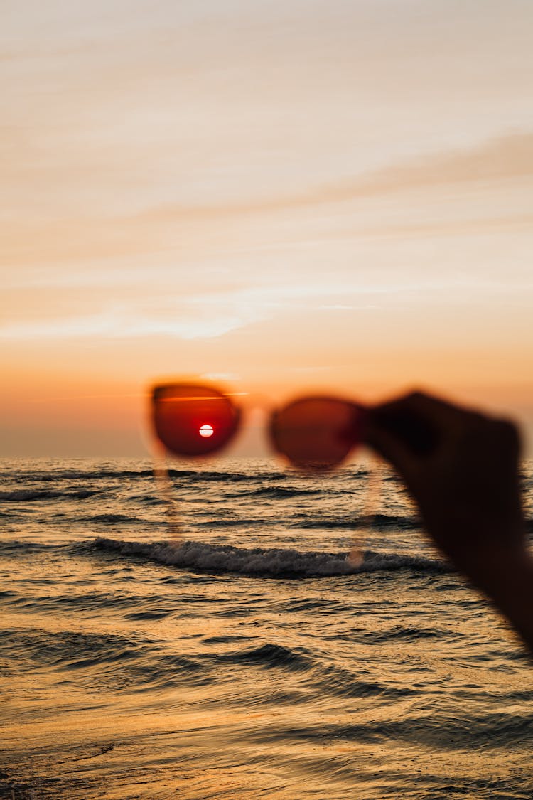 Silhouette Of A Hand Holding Sunglasses To The Sunset Over Ocean Waves