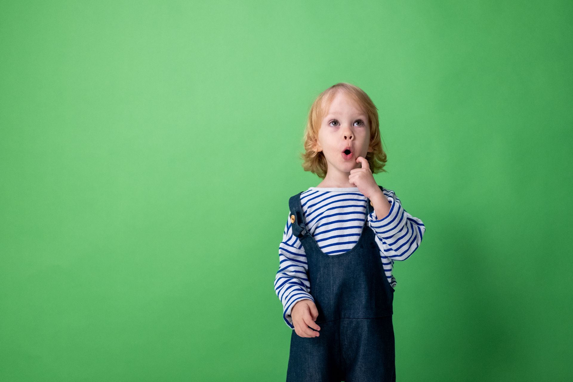 Cute child in overalls and striped shirt showing surprise against a green background in a studio setting.