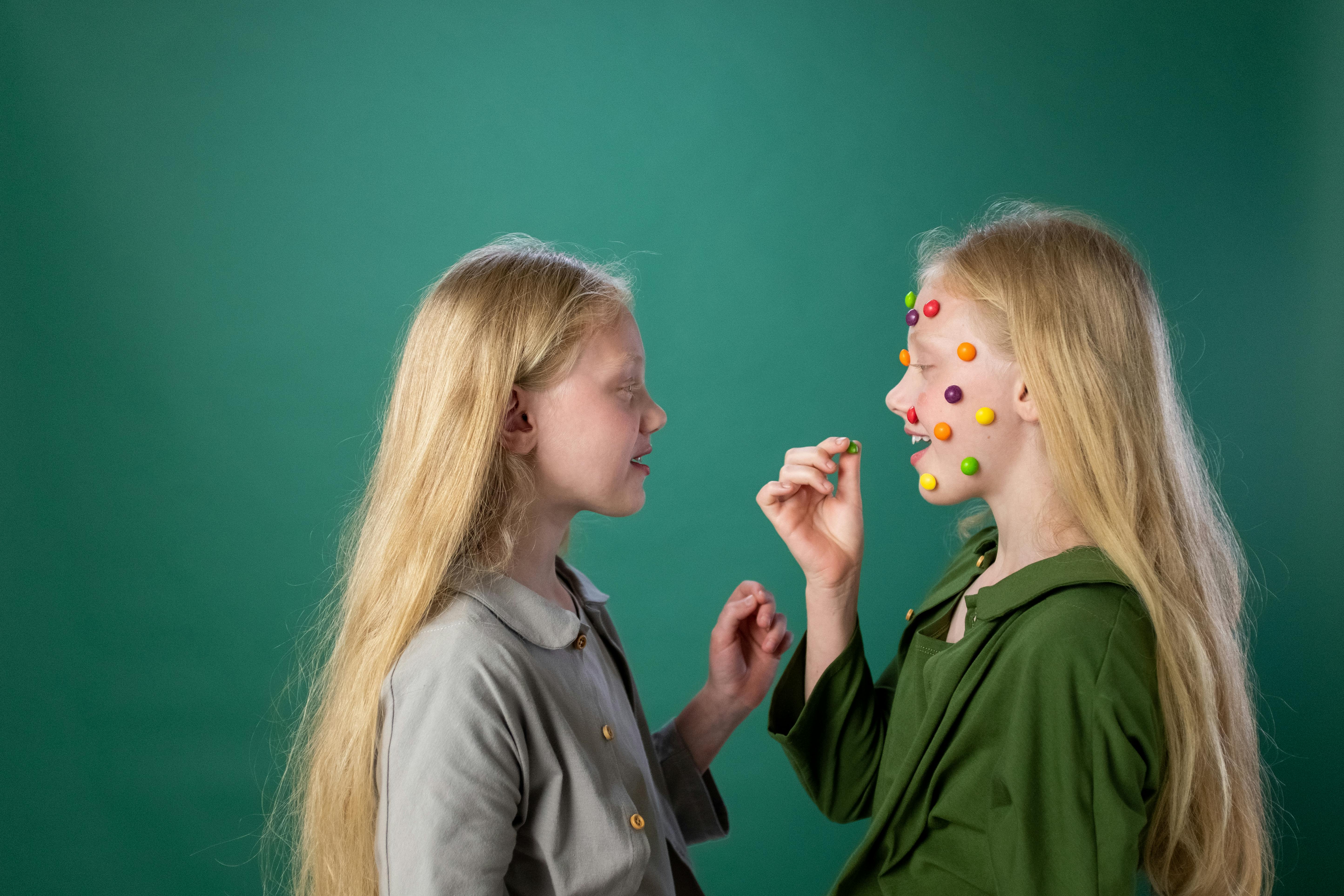 两个留着长发的年轻女孩在室内互相开玩笑，往对方脸上贴色彩缤纷的糖果贴纸