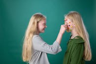 两个金发姐妹贴着彩色脸贴纸，在绿色背景前享受着有趣的时光