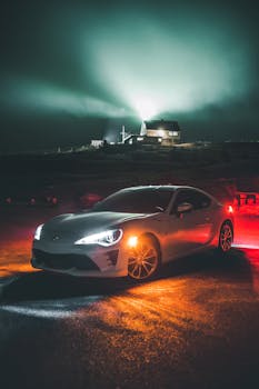 A sleek luxury car on a rural road is highlighted by an illuminated house in the distance at night.