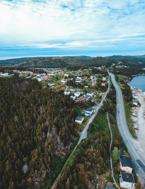 Aerial view of narrow roadway between mounts with greenery woods and village near ocean under bright cloudy sky